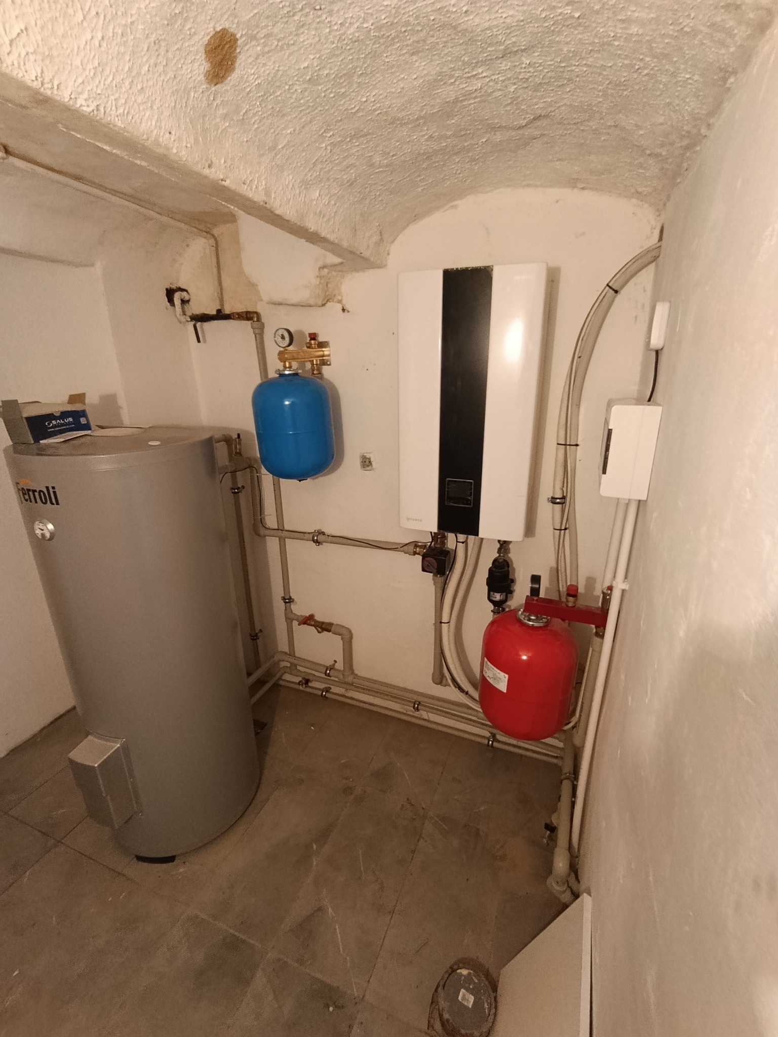 Instalacja grzewcza w piwnicy z kotłem, zasobnikiem Ferroli, naczyniami przeponowymi i rurami. Sufit łukowy.