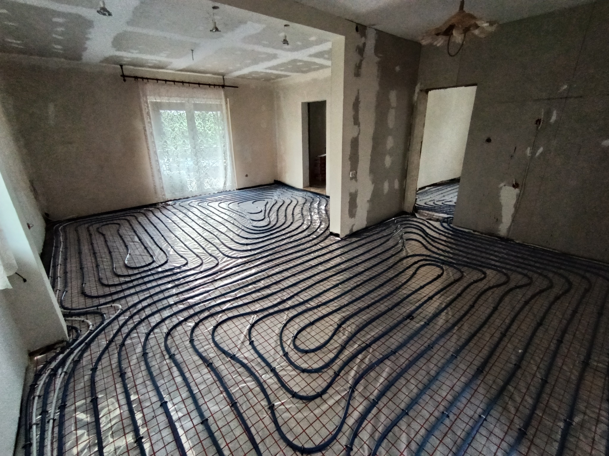 Rozłożona instalacja ogrzewania podłogowego w surowym wnętrzu. Widoczne rury na folii, przygotowane do zalania posadzką. W tle okno i surowe ściany.