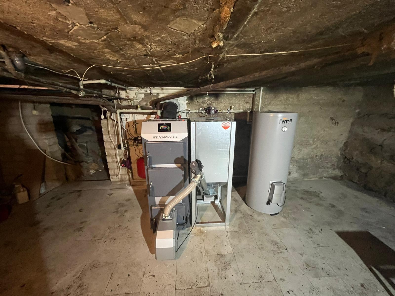 Piwnica z zainstalowanym kotłem na paliwo stałe marki Stalmark i zasobnikiem ciepłej wody Ferroli, widoczne rury instalacyjne.