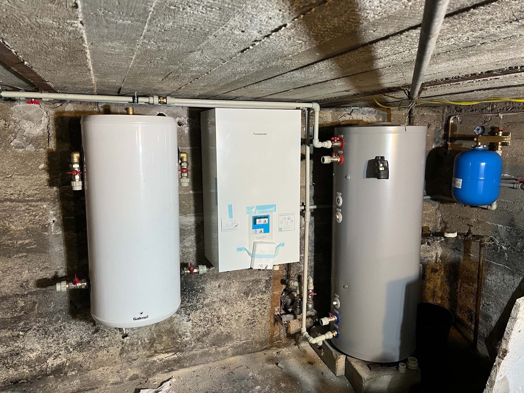 Zestaw urządzeń grzewczych: biały zbiornik Galmet, pompa ciepła Panasonic i szary zbiornik, zainstalowane w pomieszczeniu z surowym betonowym stropem.