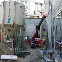 Miniaturowy żuraw samojezdny na placu budowy, podnosi szybę okienną, w tle silosy z zaprawą i budynek w stanie surowym.