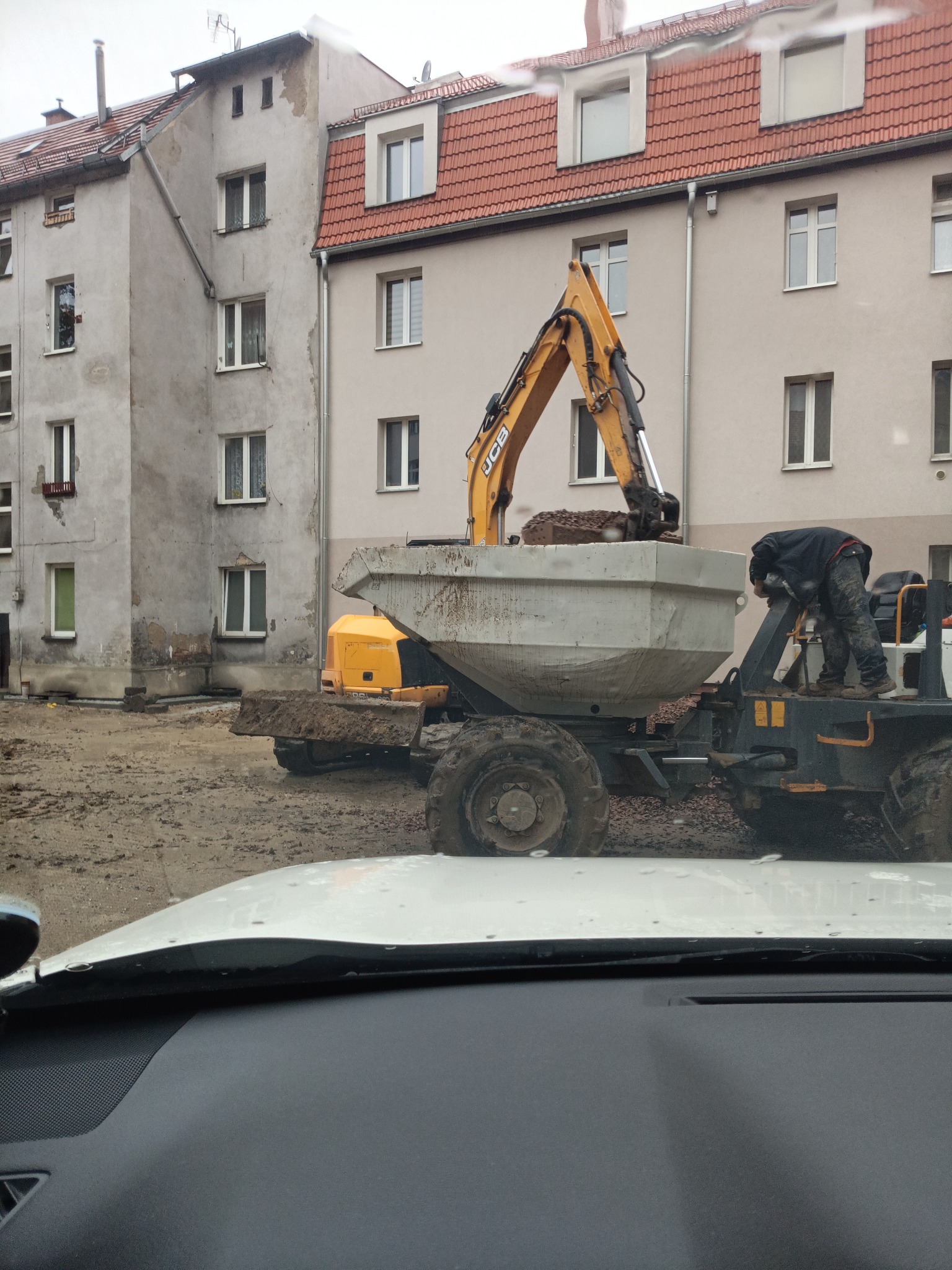Żółta koparka JCB ładuje ziemię do wywrotki na terenie budowy, w tle budynki mieszkalne z czerwonymi dachami, widok z wnętrza samochodu.