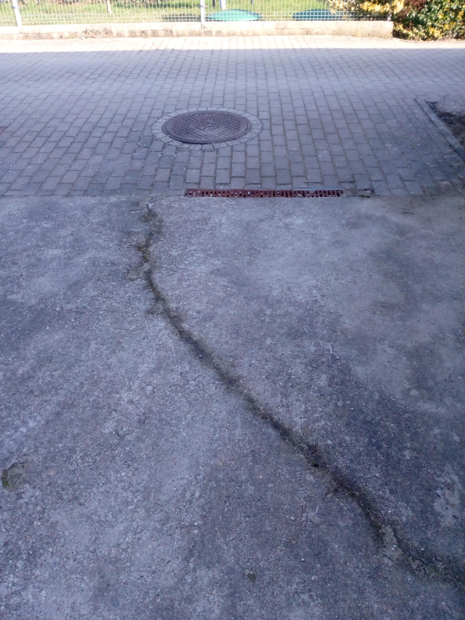 Szara, popękana betonowa nawierzchnia z widoczną szczeliną biegnącą ukośnie, przechodząca w kostkę brukową z okrągłym włazem kanalizacyjnym i kratką ściekową w tle.