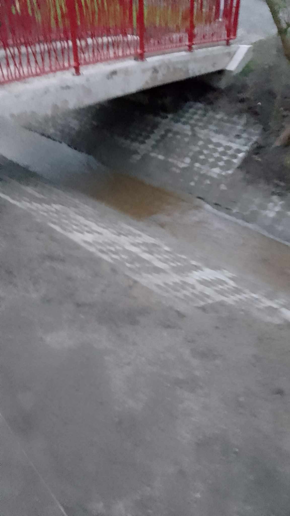 Ujęcie z dołu na betonowy mostek z czerwonymi barierkami, pod którym widać fragment ułożonej kostki brukowej na ziemi.