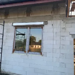 Montaż okna z brązową ramą i zamontowaną roletą zewnętrzną w surowym stanie budynku z białych bloczków, widoczne rusztowanie.