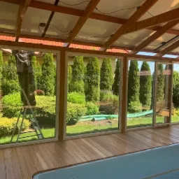 Montaż panoramicznych okien w drewnianej konstrukcji oranżerii z widokiem na zadbany ogród, pracownik na drabinie w tle.