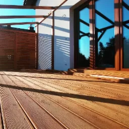 Drewniany taras z zadaszeniem w nowoczesnym stylu, widoczne duże okno i ażurowe przesłony, oświetlone popołudniowym słońcem.