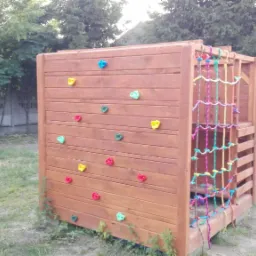 Drewniany plac zabaw dla dzieci z kolorowymi uchwytami do wspinaczki i siatką sznurkową na boku, ustawiony na trawniku w ogrodzie.