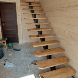 Drewniane schody wspornikowe z czarną metalową konstrukcją w surowym wnętrzu z boazerią, widoczne narzędzia i materiały budowlane na podłodze.