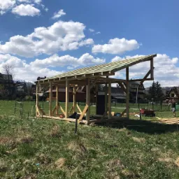 Szkielet drewnianej konstrukcji z częściowo ułożonym dachem na tle zielonej trawy, błękitnego nieba i zabudowań.