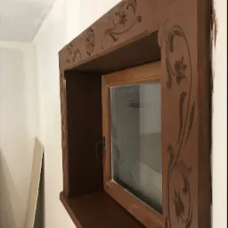 Drewniane okno z dekoracyjnymi rzeźbieniami w brązowej ramie, zamontowane w białej ścianie, widoczne z boku.