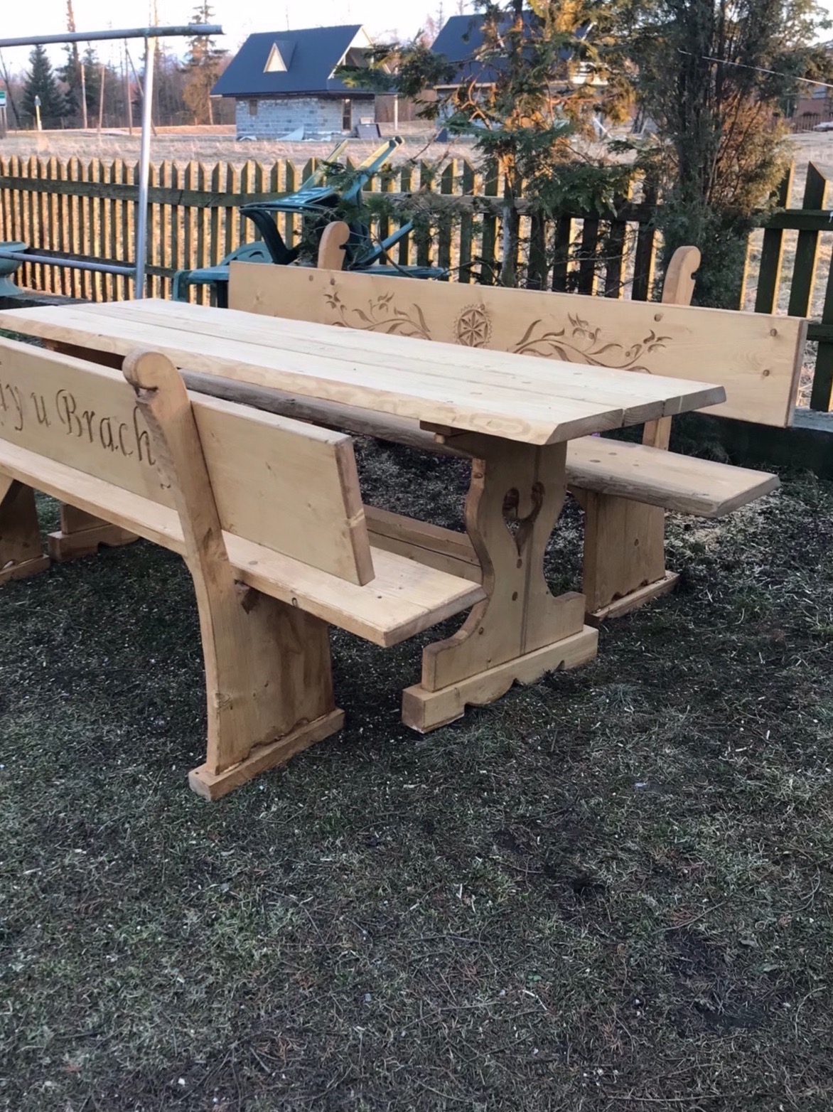 Drewniany stół z ławkami w ogrodzie, z dekoracyjnymi wycięciami i napisem 'My u Bracha' na jednej z ławek, na tle ogrodzenia i domów.