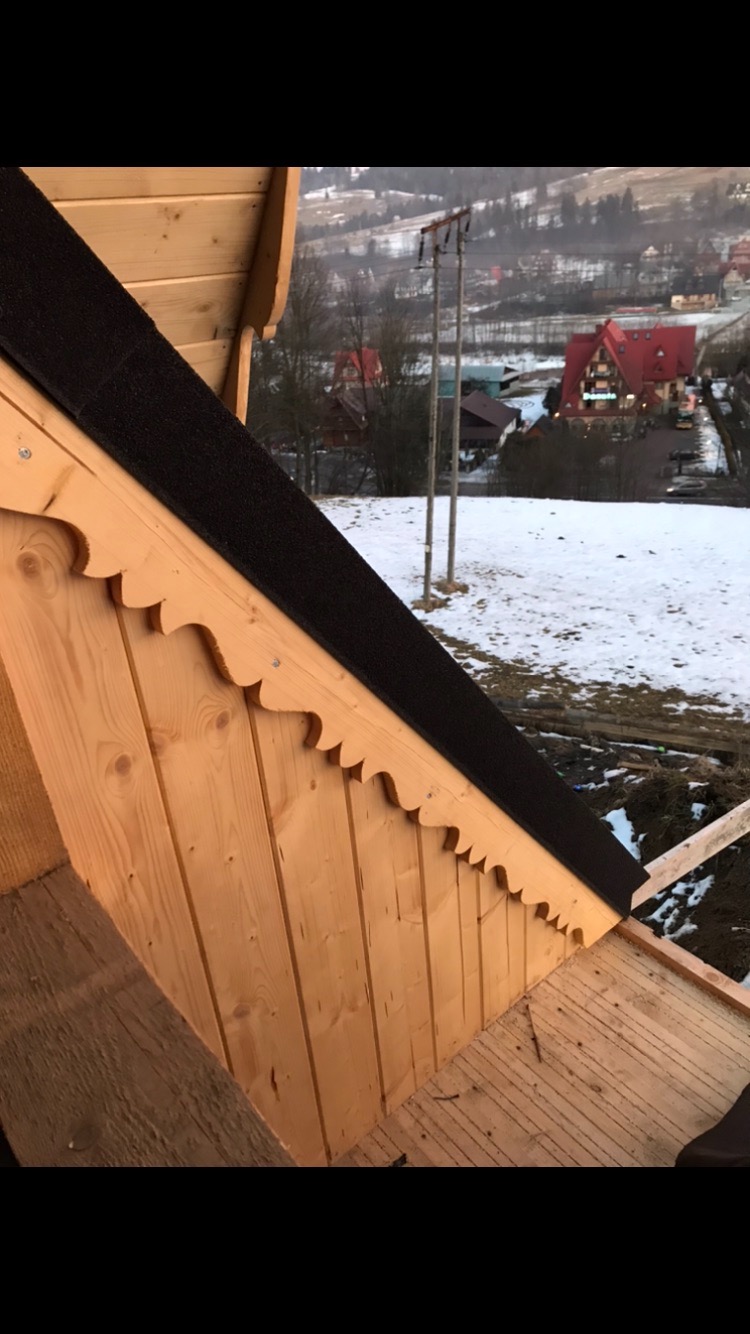 Fragment konstrukcji drewnianej dachu z dekoracyjnym wykończeniem krawędzi, widok z wnętrza, w tle ośnieżony krajobraz górski.