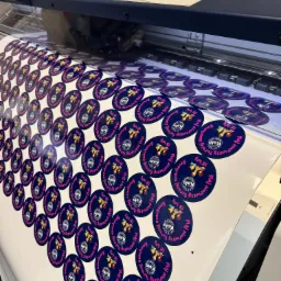 Arkusz z okrągłymi, granatowymi etykietami z różowym napisem 'Mój pierwszy turniej szachowy 15 luty' i logo Koło Materace, wychodzący z drukarki przemysłowej.