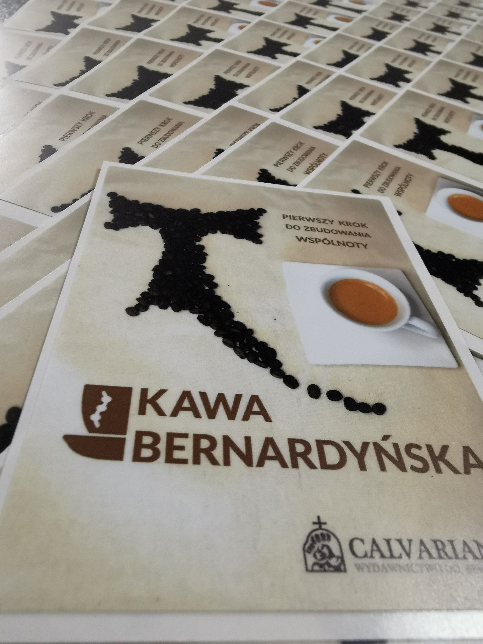 Stos naklejek z grafiką kawy bernardyńskiej, z motywem ziaren kawy ułożonych w kształt litery i filiżanki espresso, z napisem 'Pierwszy krok do zbudowania wspólnoty', tło białe.