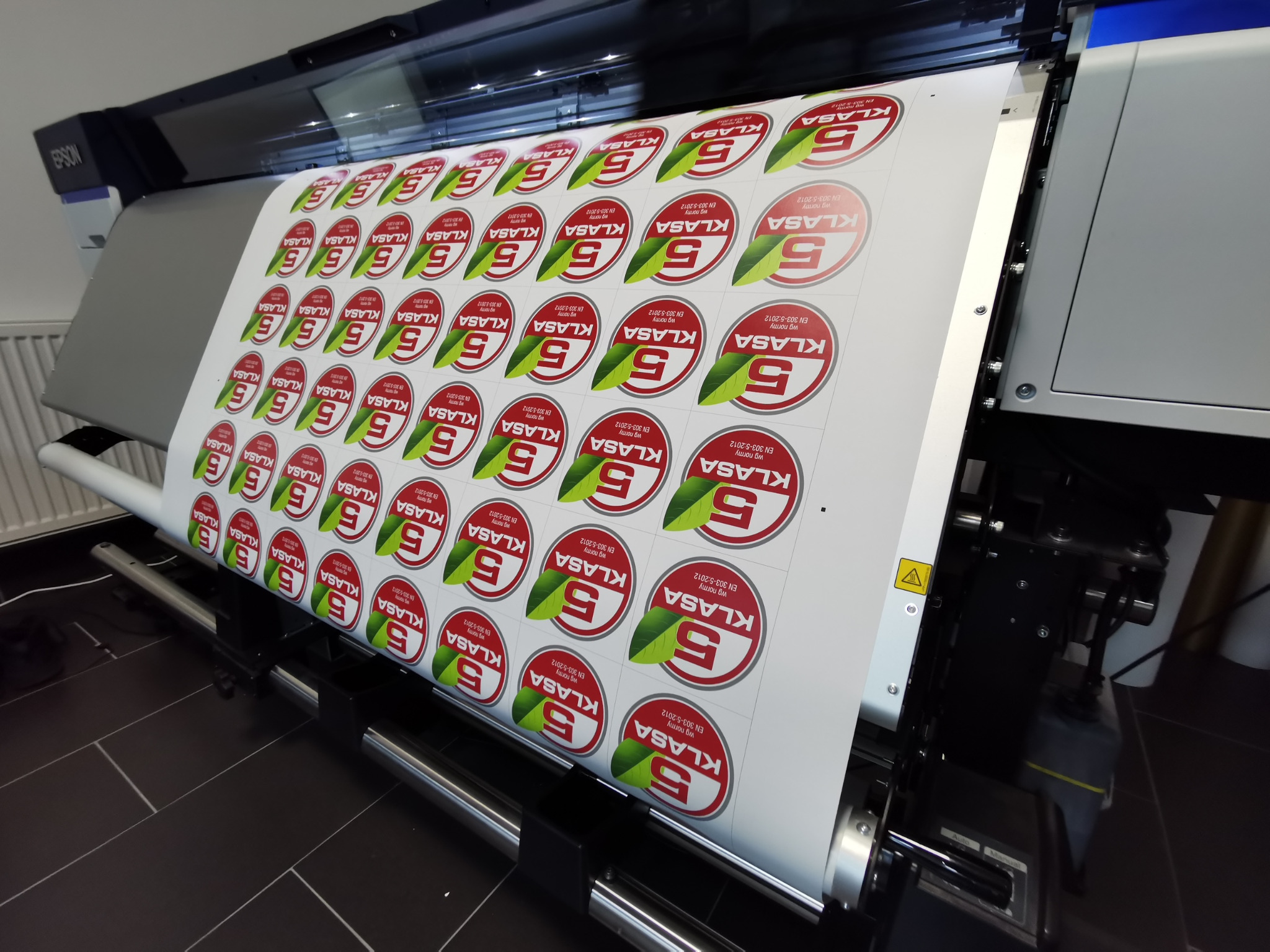Drukarka wielkoformatowa Epson drukuje arkusz z okrągłymi etykietami z zielonym liściem i napisem KLASA, zgodnie z normą EN 303-5:2012.