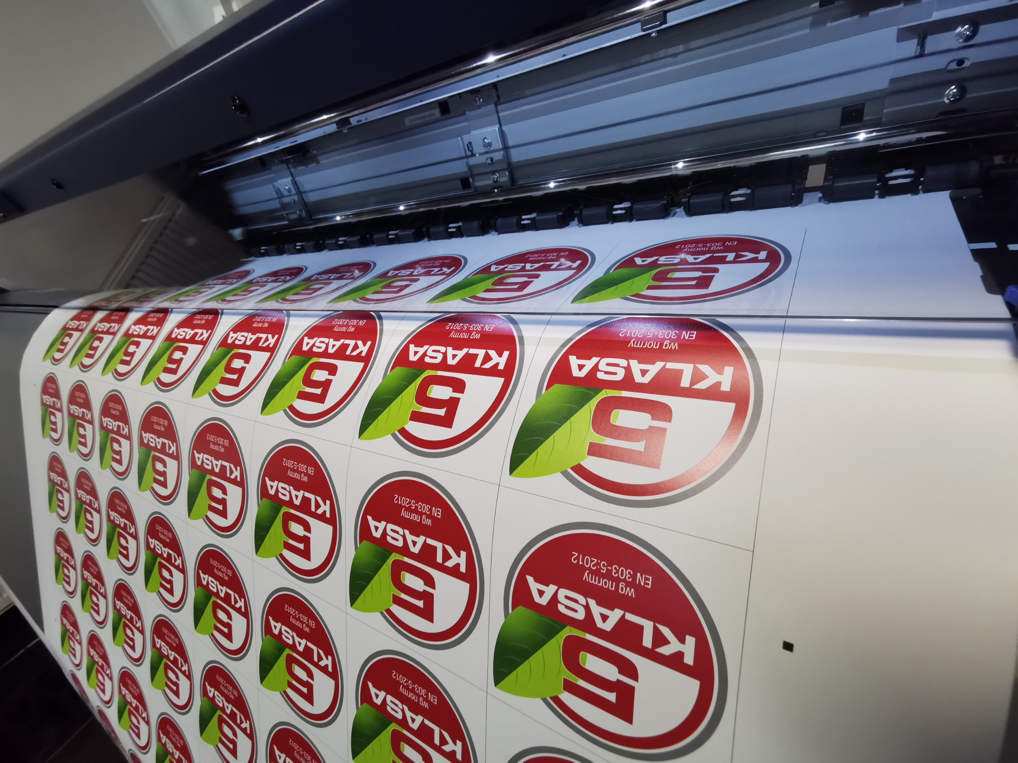 Drukarka atramentowa w trakcie drukowania arkusza z powtarzalnymi, okrągłymi etykietami z czerwoną obwódką, białym tłem, zielonym liściem i napisem 'KLASA', zgodnymi z normą EN 303-5:2012.