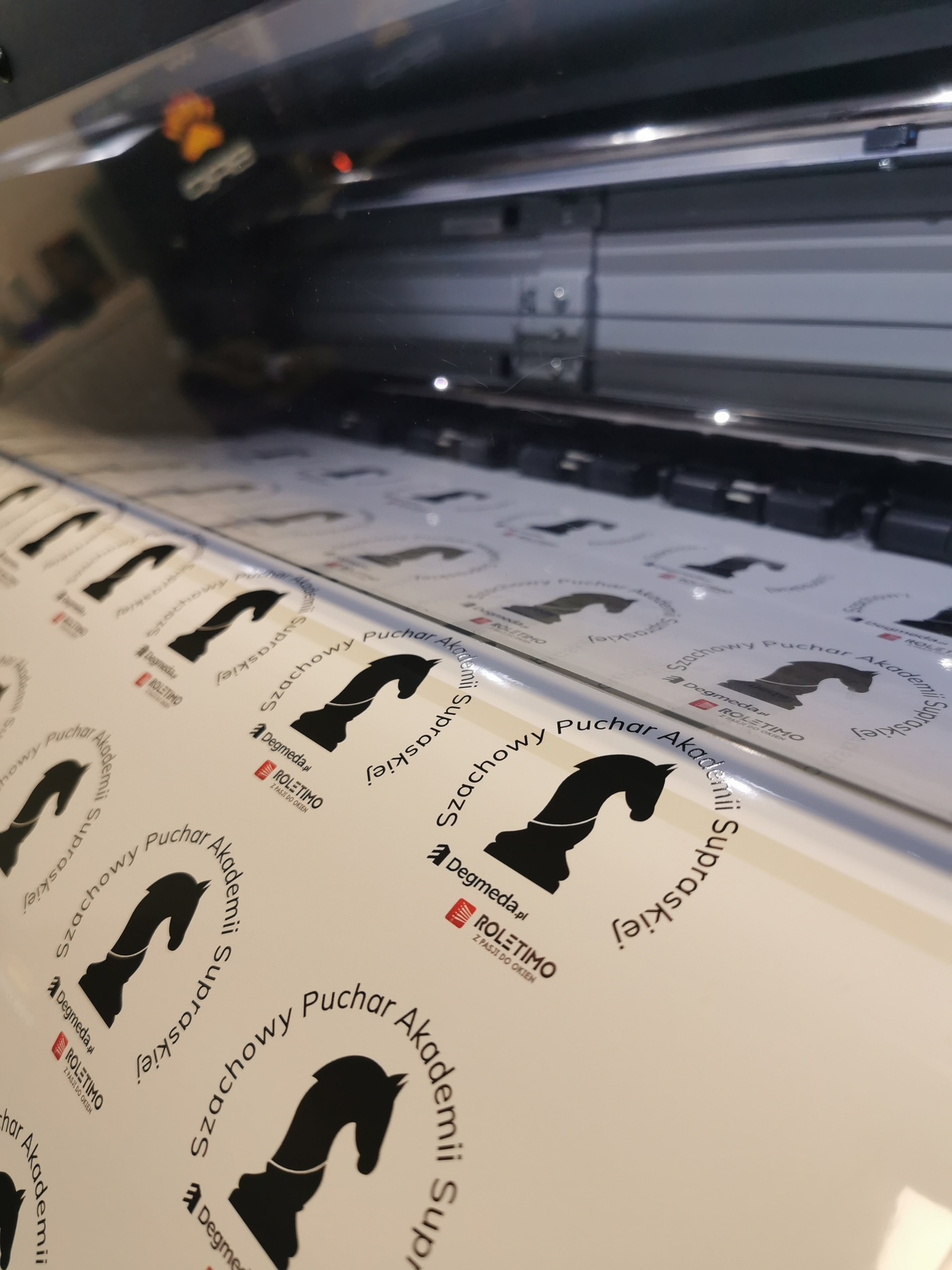 Proces drukowania naklejek z logo firmy Degmeda.pl i Roletimo, przedstawiających szachowego konia z napisem 'Szachowy Puchar Akademii Supraskiej', widoczne wewnątrz maszyny drukarskiej.
