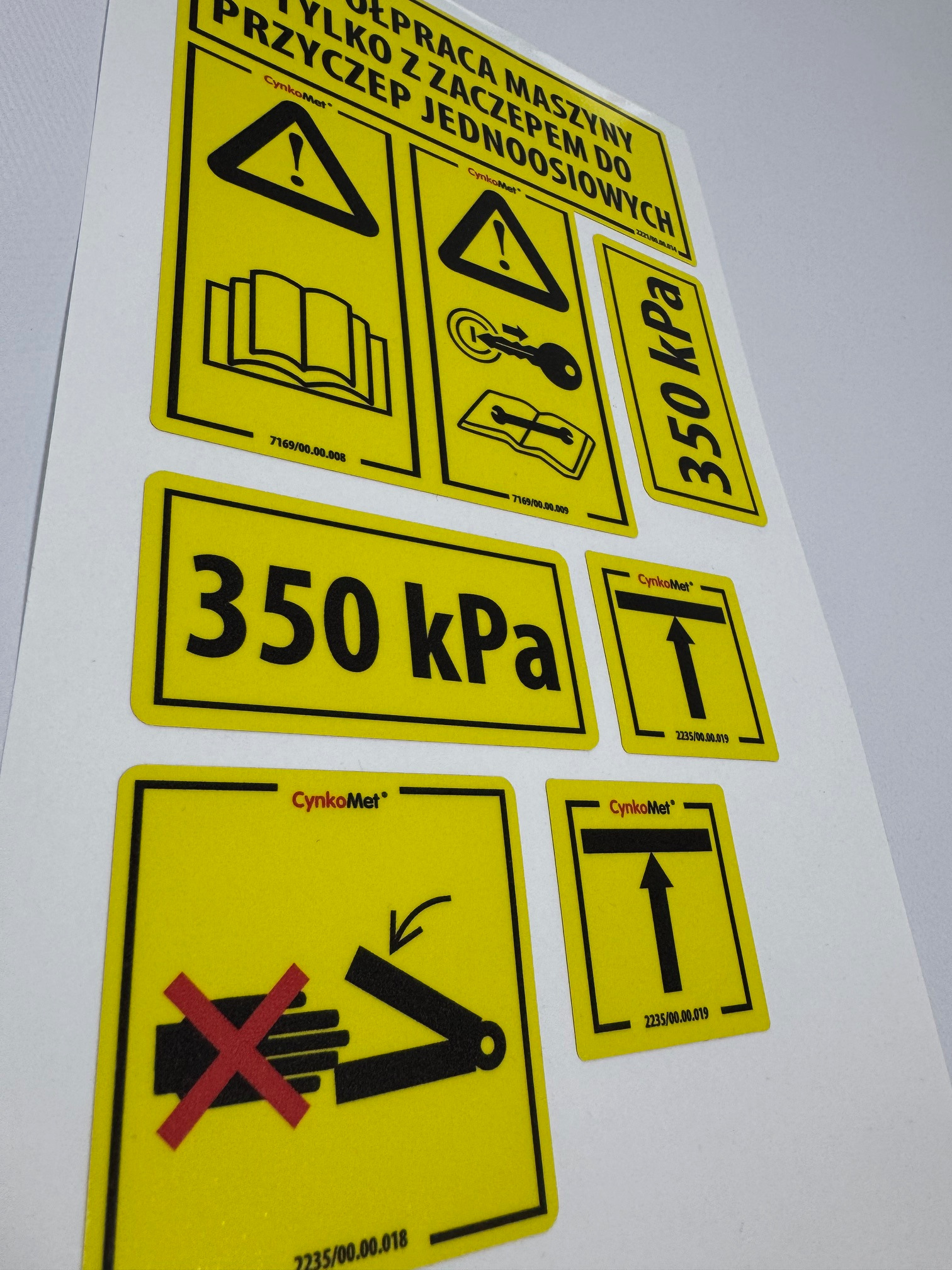 Żółte etykiety ostrzegawcze i informacyjne z czarnymi symbolami i tekstem, w tym ostrzeżenie o ciśnieniu 350 kPa i potencjalnym zagrożeniu dla rąk, naklejone na białym tle.