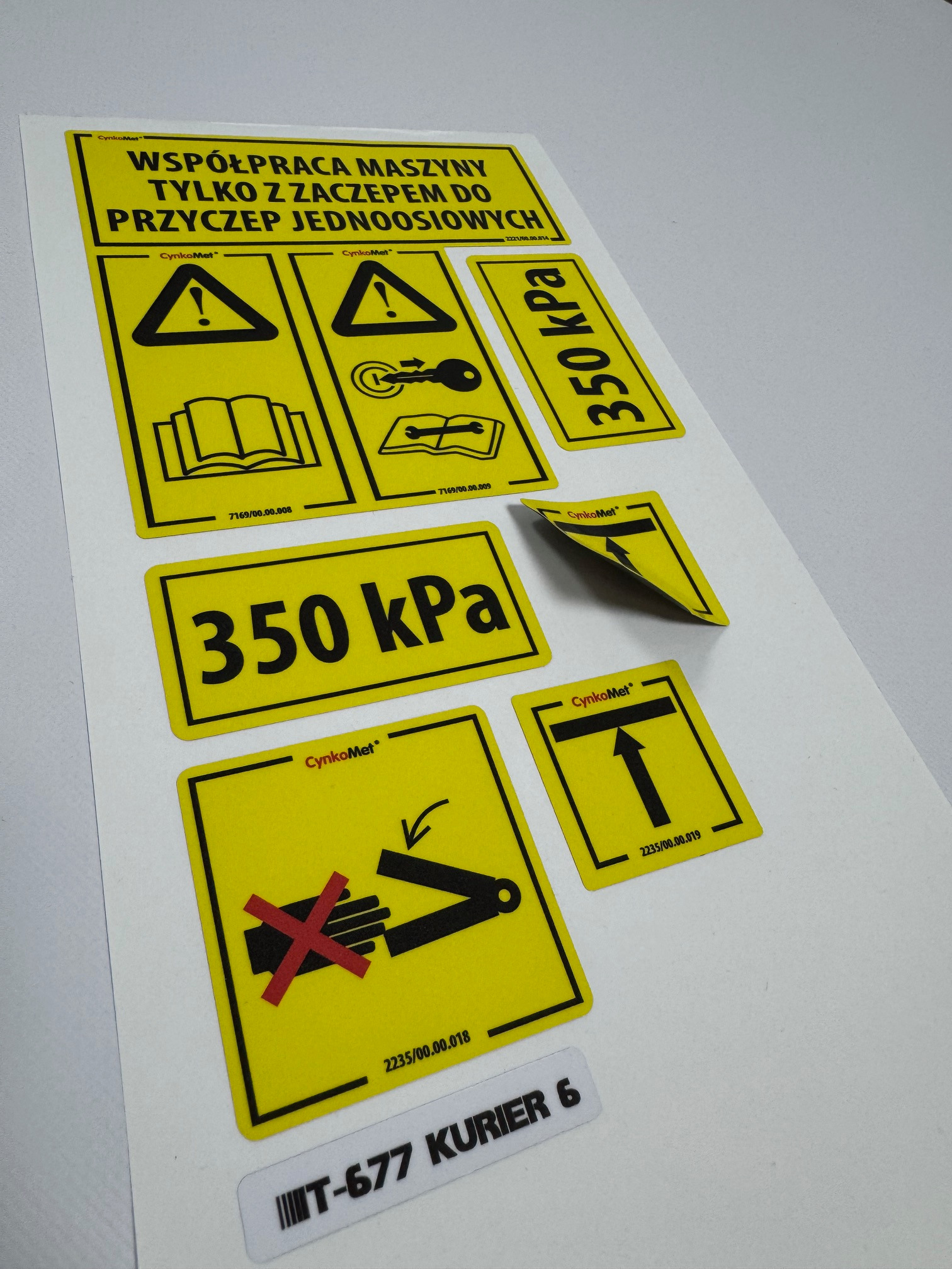 Arkusz z żółtymi znakami ostrzegawczymi i informacyjnymi, w tym symbole dotyczące bezpieczeństwa, instrukcji obsługi, ciśnienia 350 kPa oraz sposobu transportu, z widocznym odklejającym się rogiem...