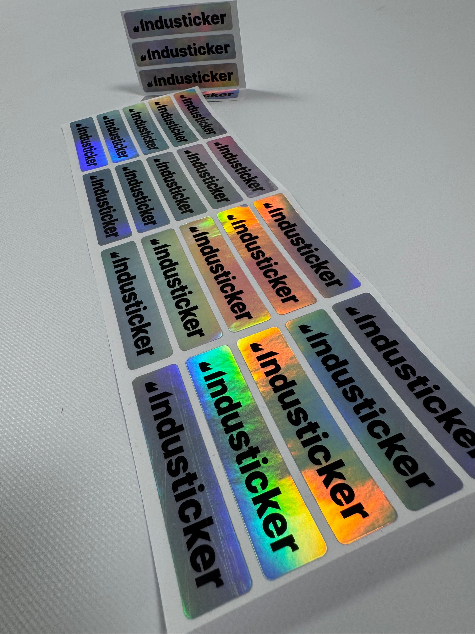 Arkusze z prostokątnymi, holograficznymi etykietami z czarnym napisem 'Industicker', ułożone pod kątem na jasnym tle.