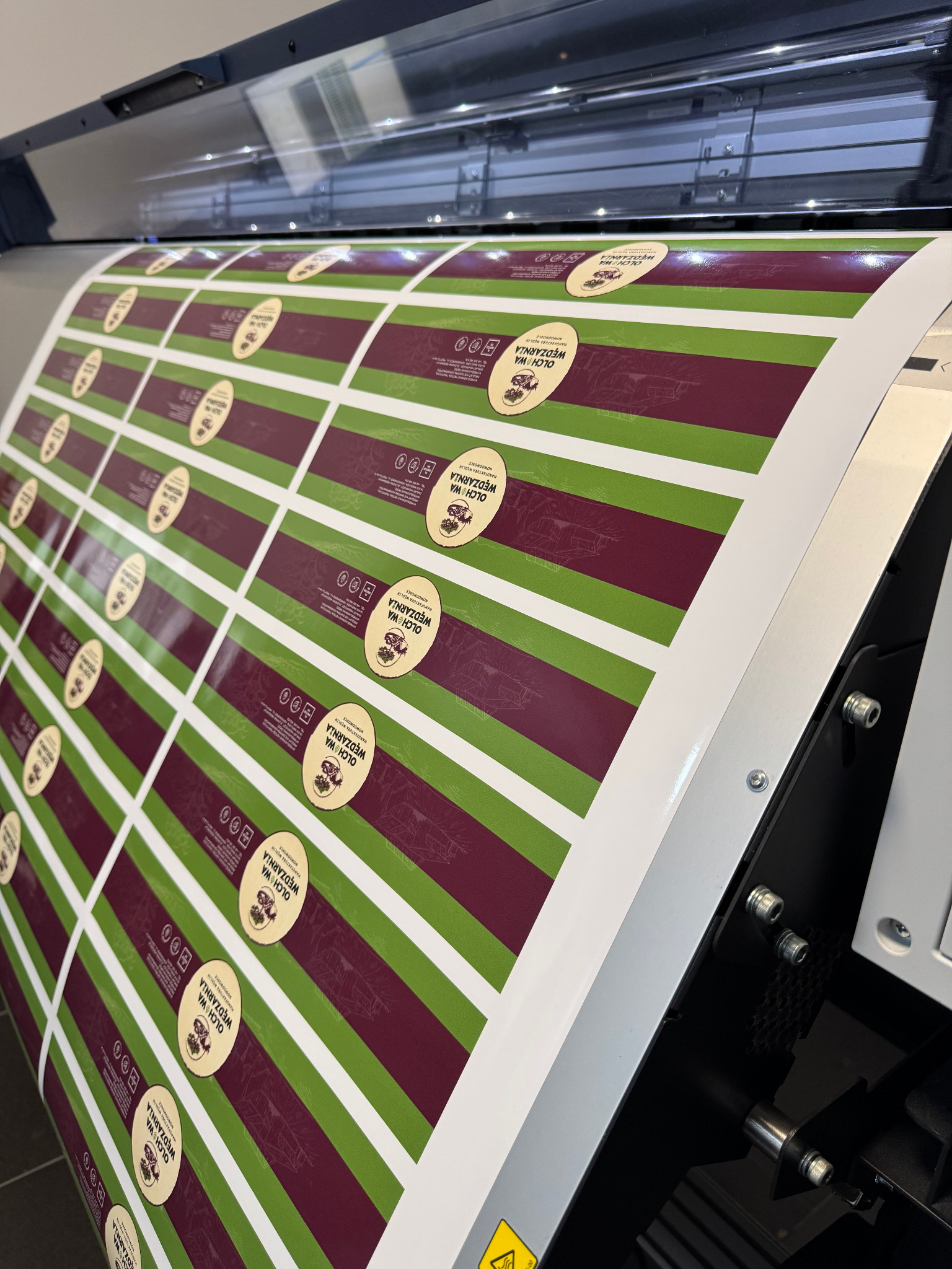 Arkusz z powtarzalnymi etykietami produktowymi 'Olchowa Wędzarnia' z grafiką wędzonego domu, w trakcie cięcia na ploterze tnącym.