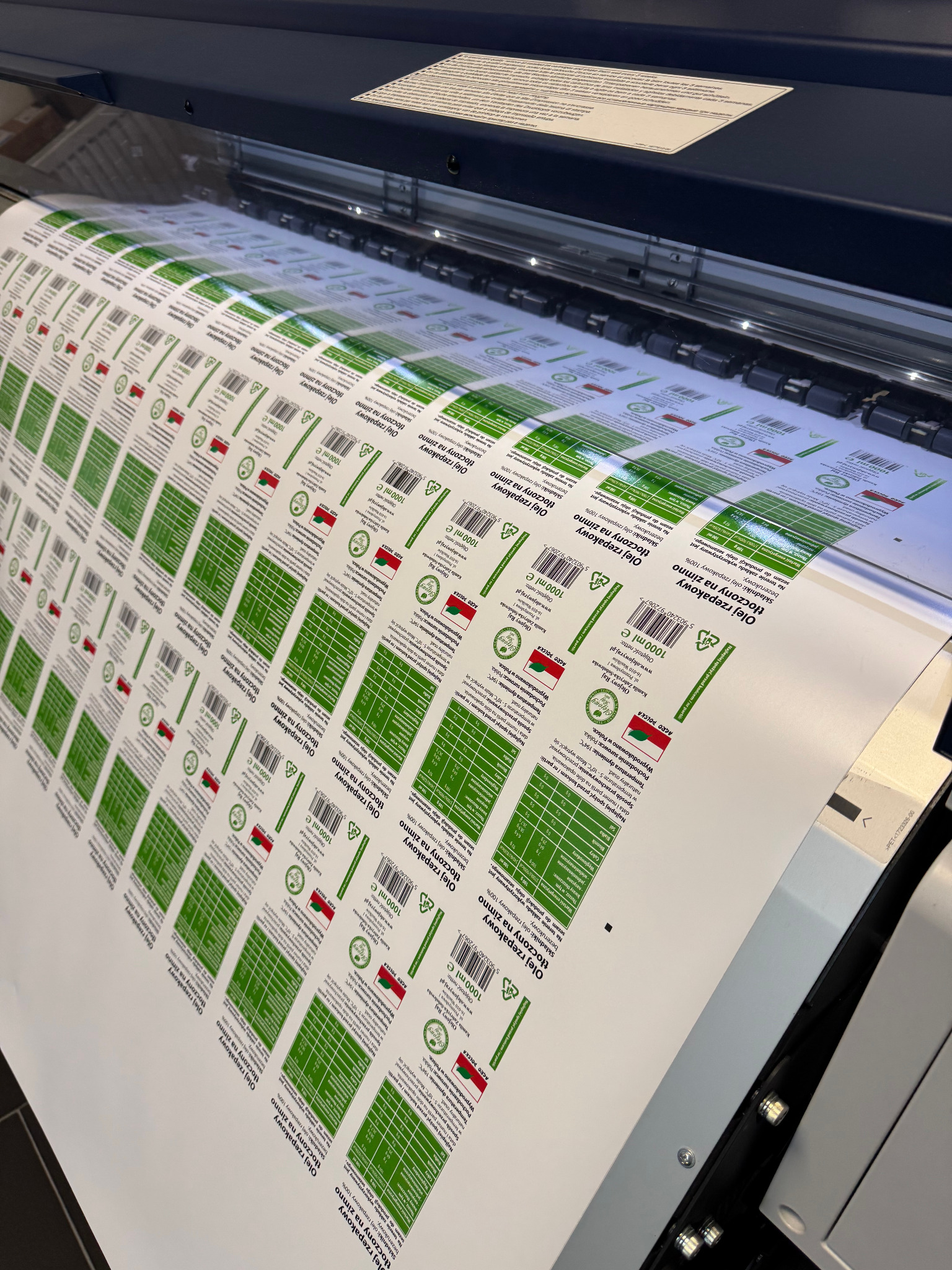 Drukarka przemysłowa drukuje arkusz z powtarzalnymi etykietami na butelki oleju rzepakowego tłoczonego na zimno, z widocznymi danymi o wartościach odżywczych i logiem producenta Agro Polska.