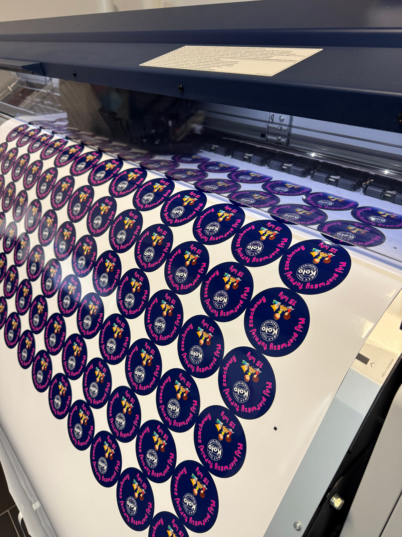 Arkusz z okrągłymi, granatowymi etykietami z różowym napisem 'Mój pierwszy turniej szachowy 15 luty' i logo Koło Materace, wychodzący z drukarki przemysłowej.