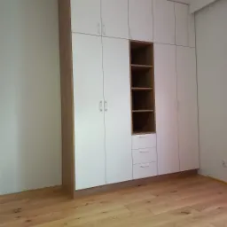 Wysoka, zabudowana szafa z białymi frontami i drewnianymi bokami, z otwartą wnęką z półkami i trzema szufladami, na drewnianej podłodze.