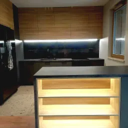 Nowoczesna kuchnia z drewnianymi szafkami górnymi, czarnym blatem i wyspą z podświetlanymi półkami, widok na okno w tle.