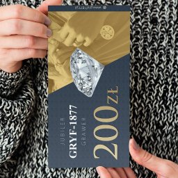 Piotr Zgorzelski - Dłonie trzymają kartę podarunkową jubilera Gryf-1877 z brylantem i kwotą 200 zł, na tle swetra o grubym splocie.