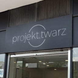 Piotr Zgorzelski - Szyld firmy 'projekt.twarz' nad wejściem do budynku, z minimalistycznym logo w postaci okręgu i napisu, na ciemnoszarym tle.