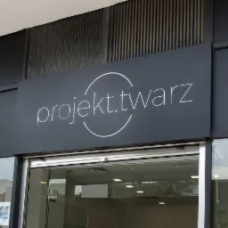 Szyld firmy 'projekt.twarz' nad wejściem do budynku, z minimalistycznym logo w postaci okręgu i napisu, na ciemnoszarym tle.