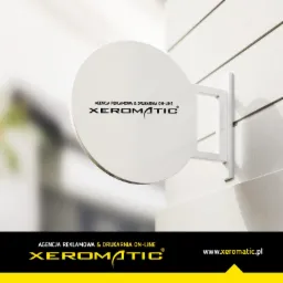 Okrągły szyld reklamowy z logo 'Xeromatic' na białej ścianie z poziomymi rowkami, prezentujący agencję reklamową i drukarnię on-line.