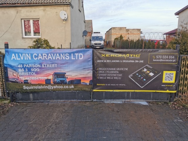 Dwa banery reklamowe zawieszone na bramie wjazdowej: jeden z ofertą sprzedaży kamperów, drugi z ofertą agencji reklamowej i drukarni online, z widocznym tabletem i kodem QR.