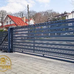 Nowoczesna, przesuwna brama wjazdowa z poziomymi panelami w kolorze grafitowym, zamontowana na posesji z kostki brukowej, widoczny dom z czerwonym dachem w tle oraz logo firmy w rogu.