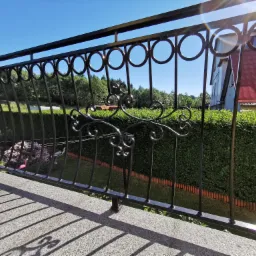 Czarna, metalowa balustrada balkonowa z dekoracyjnymi elementami w kształcie kół i ornamentów, z widokiem na zadbany ogród z żywopłotem i dom z czerwoną dachówką w tle, oświetlona jasnym...