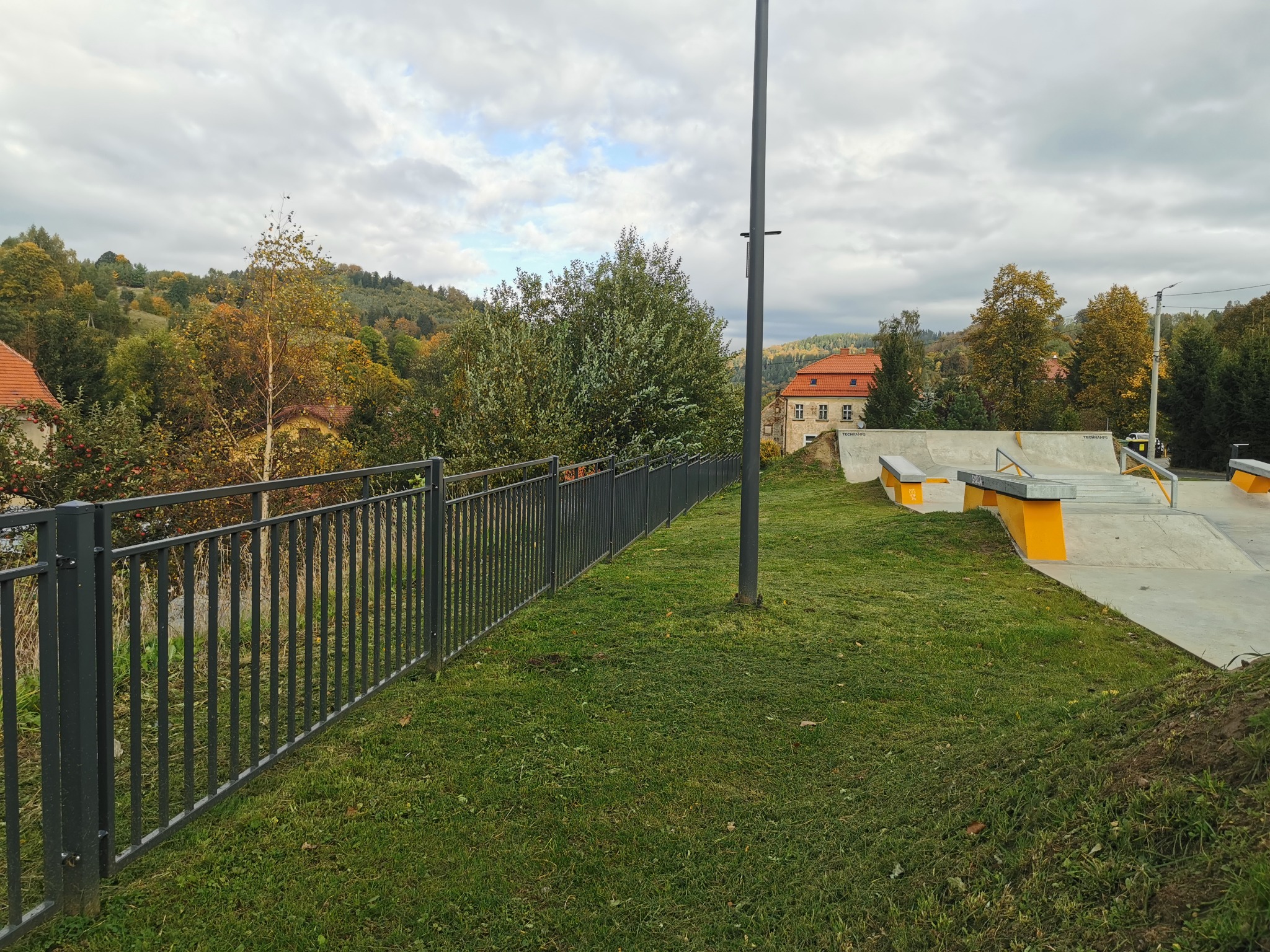 Ciemnoszare, pionowe ogrodzenie metalowe z widokiem na skatepark z żółtymi elementami, zieleń trawy i drzewa w tle pod pochmurnym niebem.