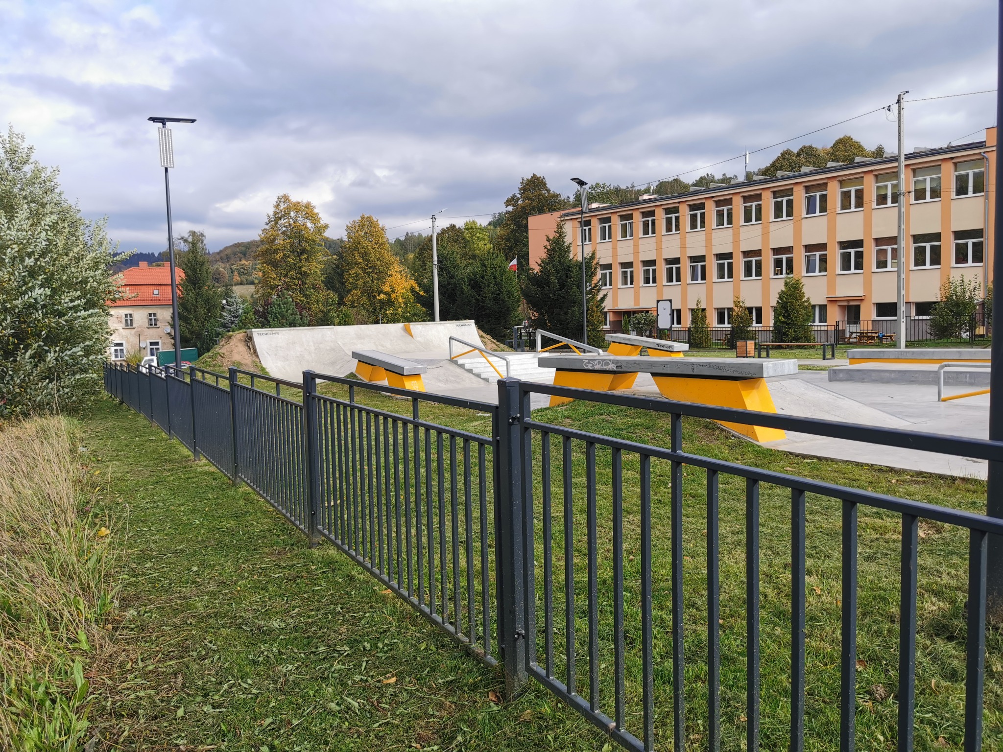 Szary metalowy płot na tle skateparku z rampami i przeszkodami w kolorze żółtym i szarym, w oddali budynek szkolny i drzewa pod pochmurnym niebem.