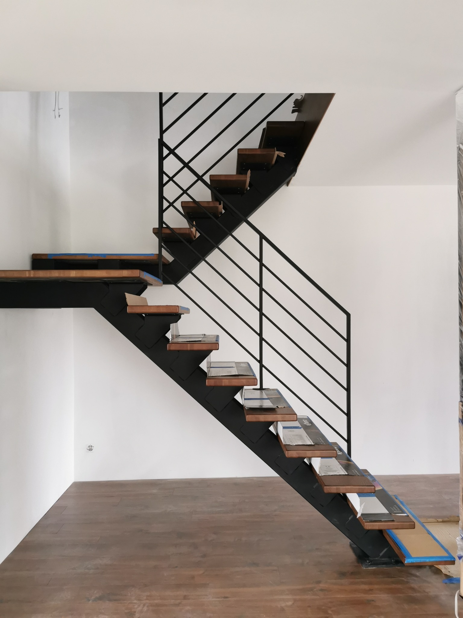 Nowoczesne schody z drewnianymi stopniami i czarną metalową konstrukcją, częściowo zabezpieczone folią i kartonem podczas montażu, widok z dołu na białą ścianę i drewnianą podłogę.