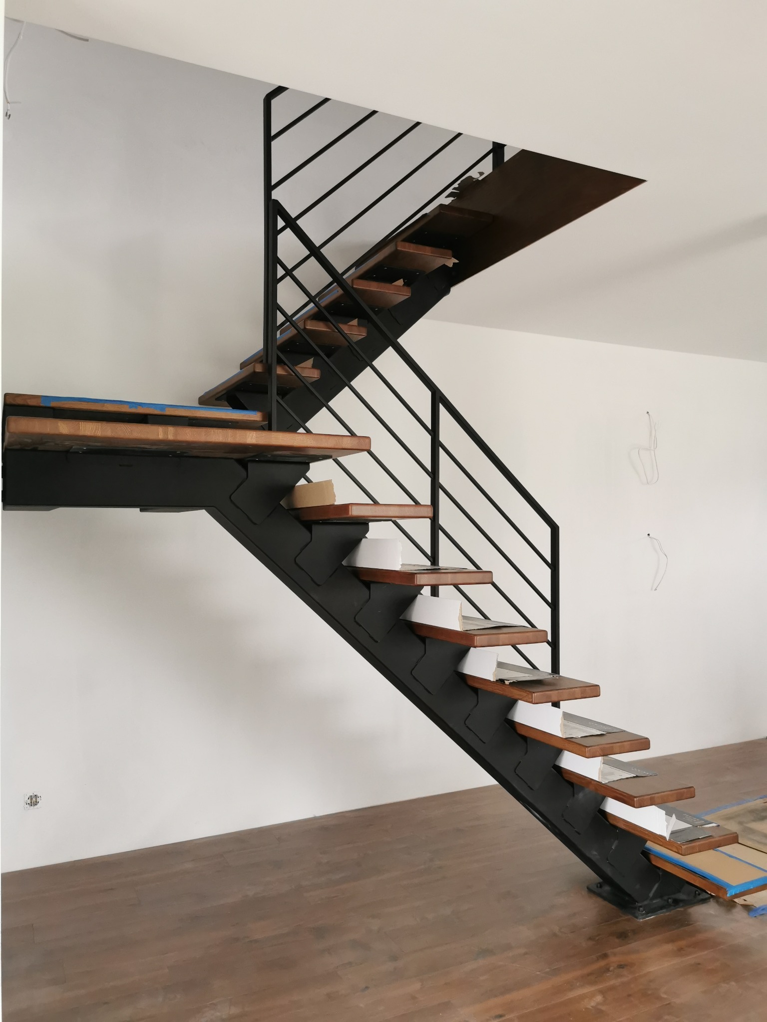 Wnętrze w trakcie wykańczania z zamontowanymi schodami metalowymi z drewnianymi stopniami, zabezpieczonymi kartonem i taśmą malarską, czarna metalowa konstrukcja i balustrada.