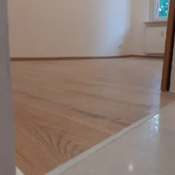 Jasny, drewnopodobny panel podłogowy ułożony w pokoju, widoczny fragment białej listwy przypodłogowej i przejście do błyszczącej, jasnej podłogi w innym pomieszczeniu.