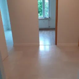 Wykończone wnętrze mieszkania z nową, jasną podłogą z płytek, widoczne białe ściany i fragment drugiego pomieszczenia z drewnianą podłogą i oknem.