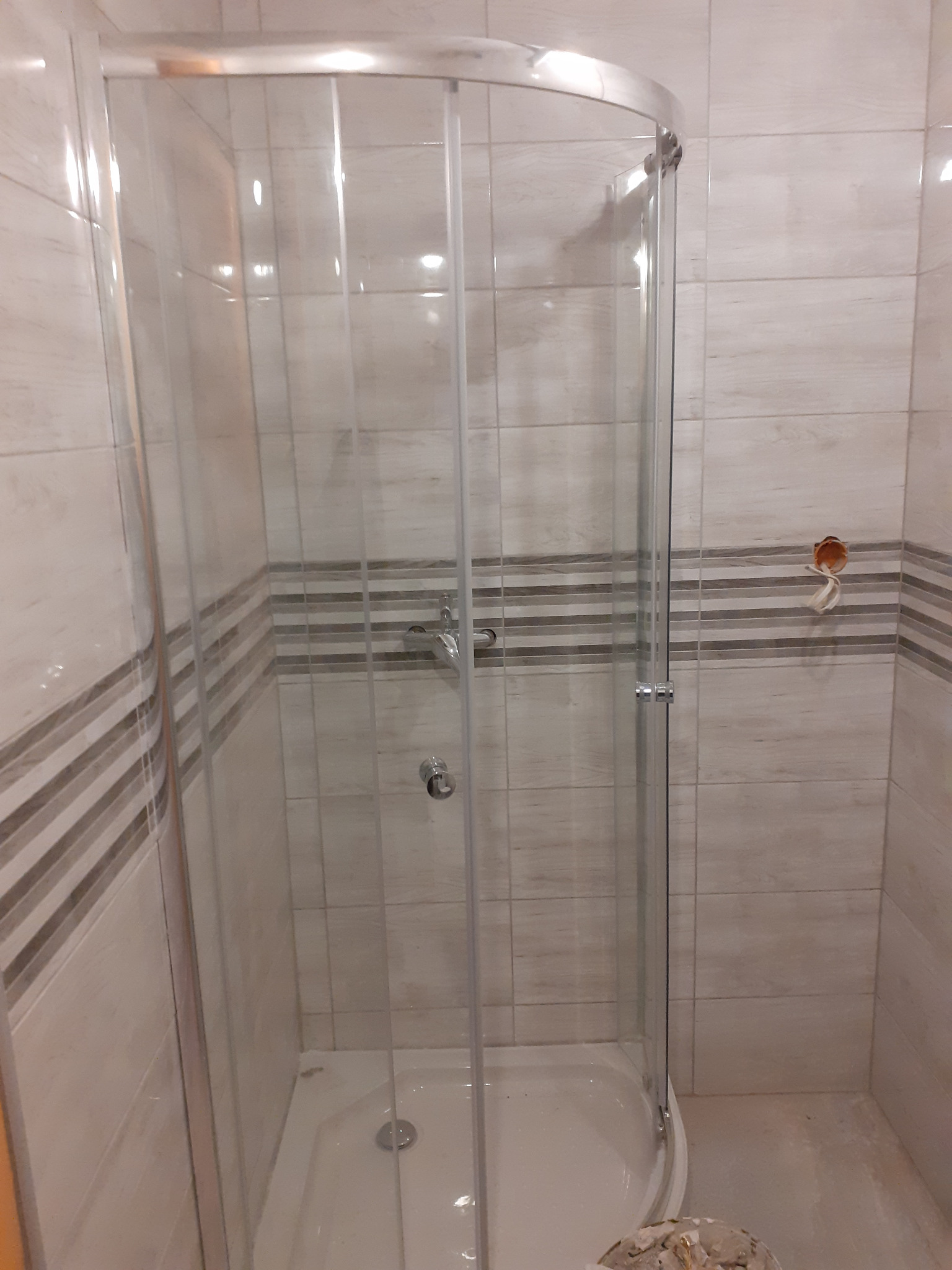 Zamontowana kabina prysznicowa z chromowanymi profilami i przezroczystymi szybami w łazience z beżowymi płytkami z dekoracyjnym pasem, widoczne niedokończone podłączenie elektryczne.