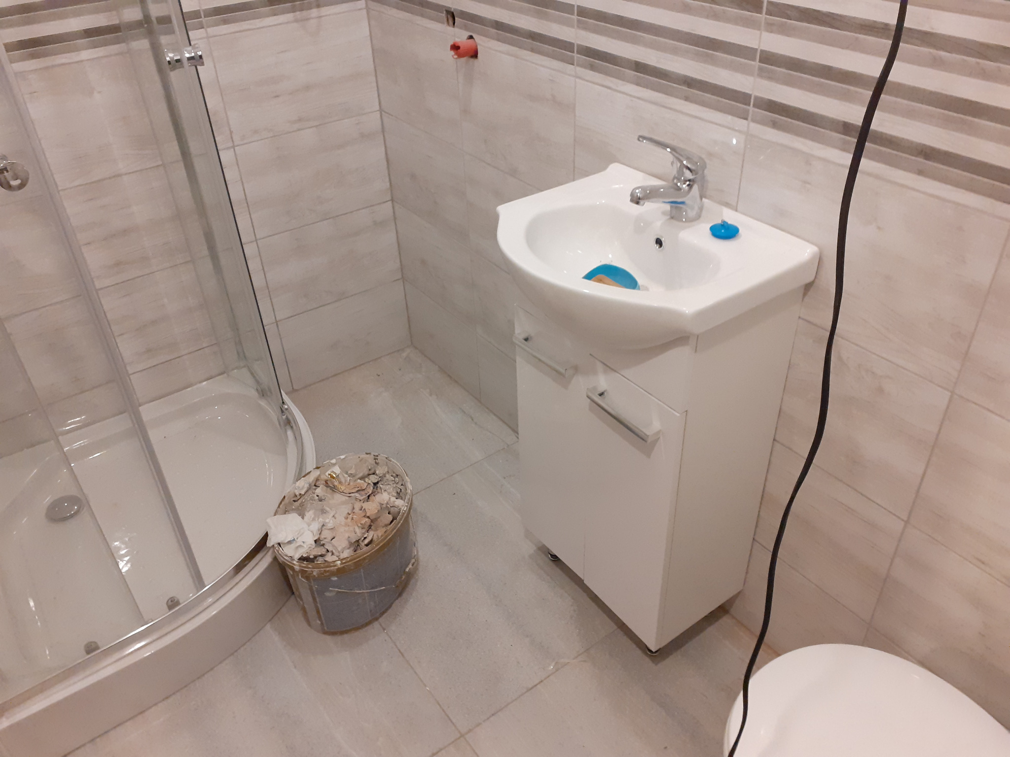 Biała umywalka z szafką, kabina prysznicowa i wiadro z resztkami po remoncie w łazience wyłożonej płytkami w odcieniach beżu i brązu.