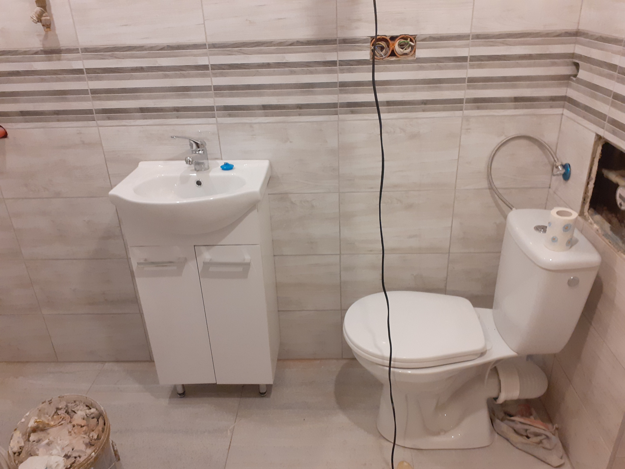 Łazienka w trakcie remontu: biała umywalka z szafką, toaleta z papierem toaletowym, ściany wyłożone jasnymi płytkami z szarymi paskami, widoczne puszki elektryczne i kabel, wiadro z zaprawą.