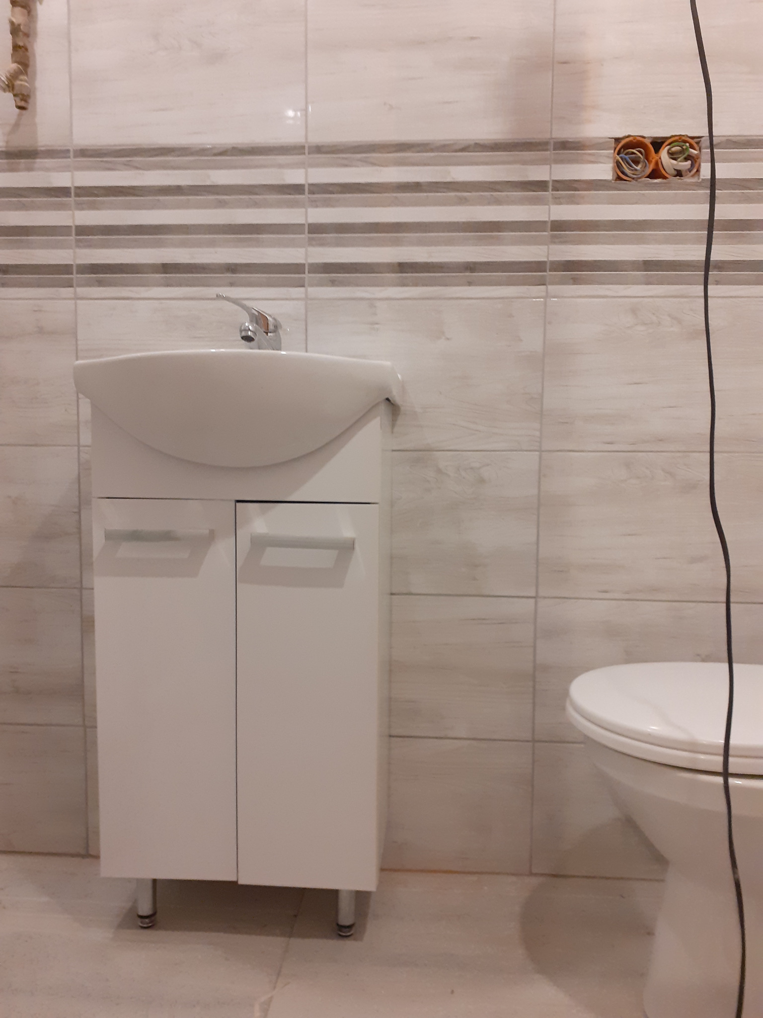 Biała szafka pod umywalkę z chromowaną baterią w łazience z beżowo-szarymi płytkami, widoczne niedokończone gniazdko elektryczne z przewodami.