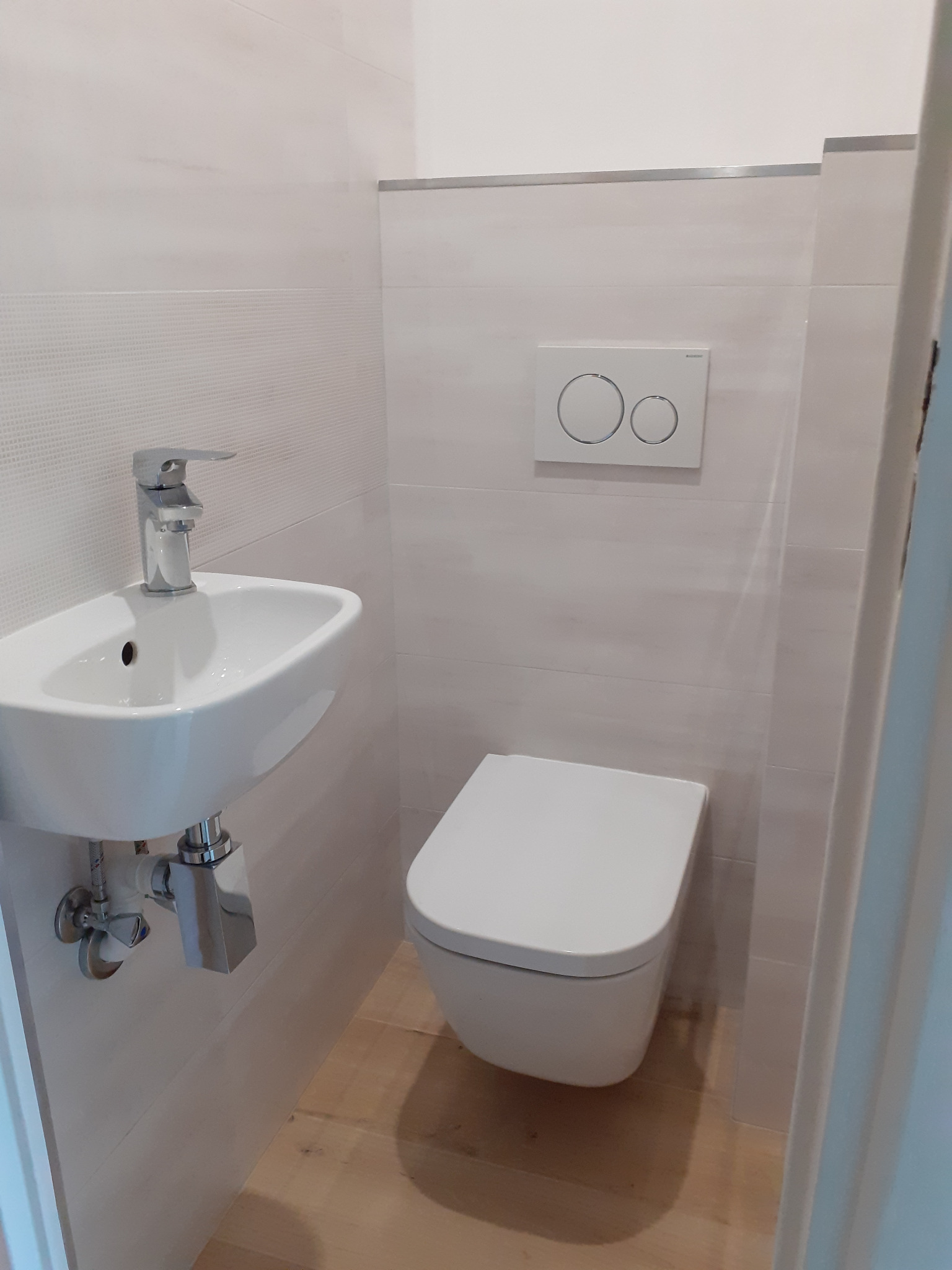 Nowoczesna, jasna łazienka z podwieszaną toaletą, umywalką narożną i płytkami imitującymi drewno na podłodze. Widoczny chromowany syfon i bateria umywalkowa.