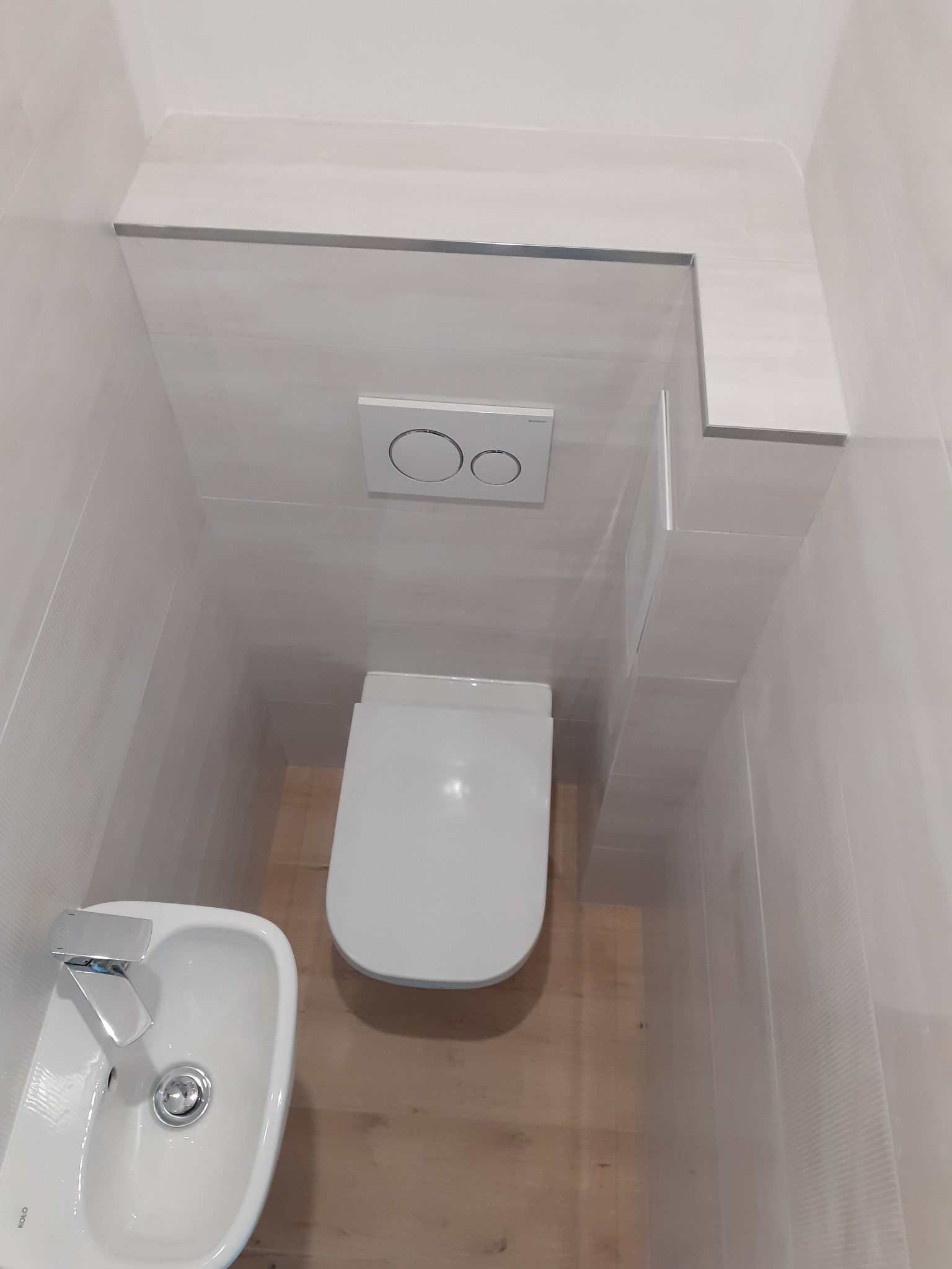 Nowoczesna, minimalistyczna toaleta z podtynkową spłuczką i małą umywalką w narożniku, wykończona płytkami w odcieniach bieli i beżu.