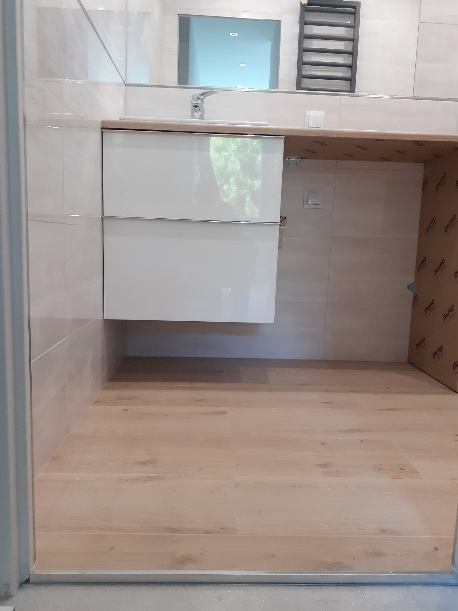 Nowoczesna łazienka z jasnymi płytkami imitującymi kamień, podwieszaną szafką z umywalką i drewnianą podłogą, widok z poziomu podłogi.